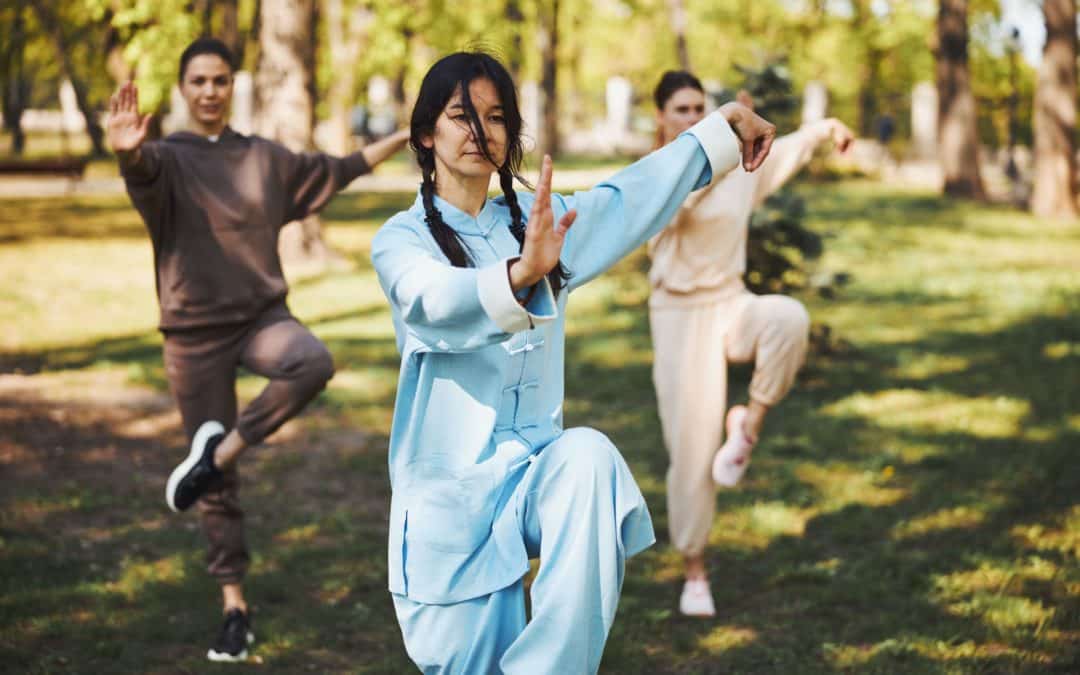 Woman martial arts master staying on right leg outdoors while showing her palm to camera with pair of students