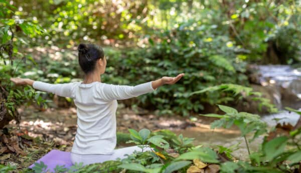 woman in white, seated with arms open practicing qigong in peaceful nature setting